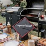 Backyard grill next to a long wooden table set with paper plates, striped napkins and a graduation cap that says Congratulations