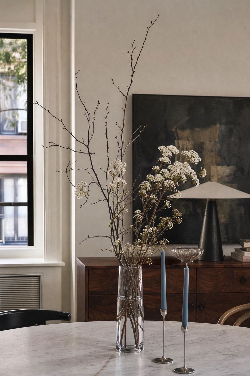 Clear glass vase with metallic branches, white florals, dusty blue taper candles, and walnut dining room details.