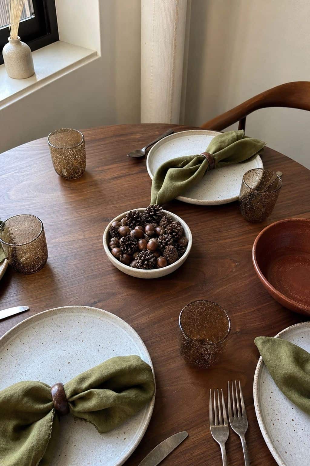 Small ceramic bowl of acorns and mini pinecones on a walnut round Thanksgiving table with olive napkins